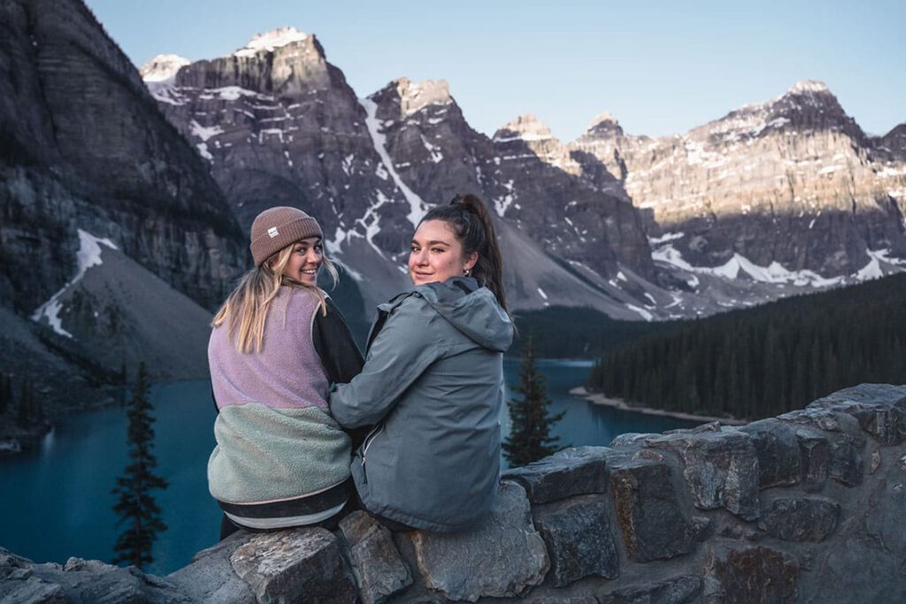 Two women sitting on a rock wall turning to look at the camera. There’s a lake and mountains in the background