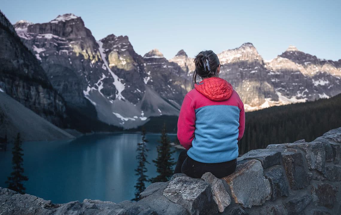 The back of a woman in a pink and blue fleece; she's sitting on a rock wall looking at a lake