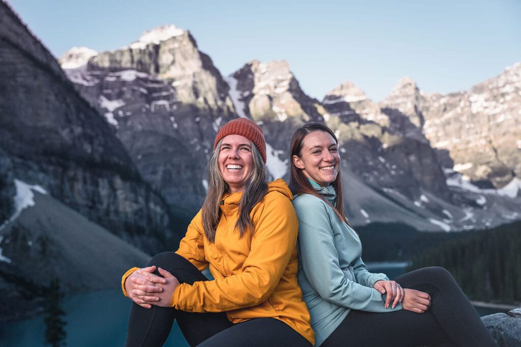 Two ladies sitting back-to-back by a lake smiling