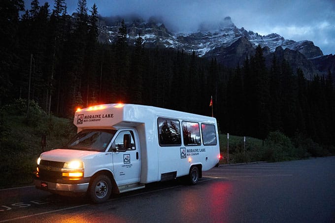A dark photo of a white Moraine Lake Bus Company shuttle in a car park with its lights on. There are trees and mountains in the backdrop