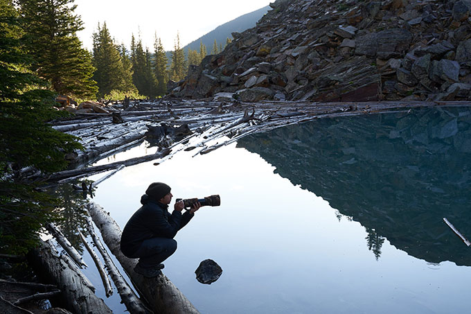 A person crouching down on a wooden log on a lake. They’re holding a large camera, about to take a photo