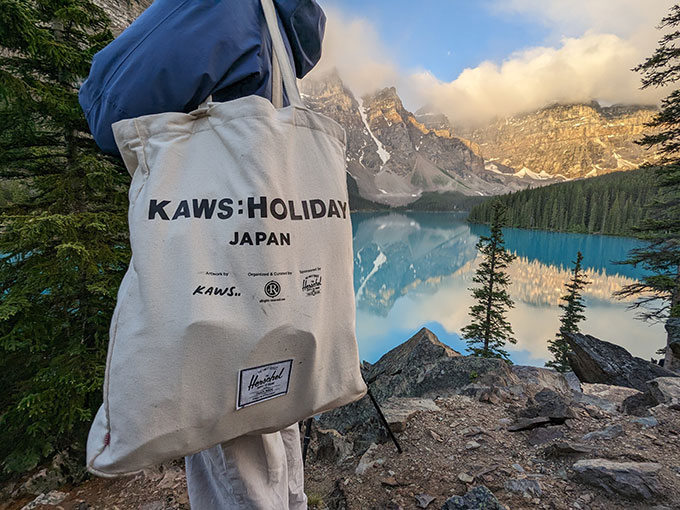 A close-up of a white tote bag that says ”˜KAWS: HOLIDAY Japan’ with a bright blue lake in the background