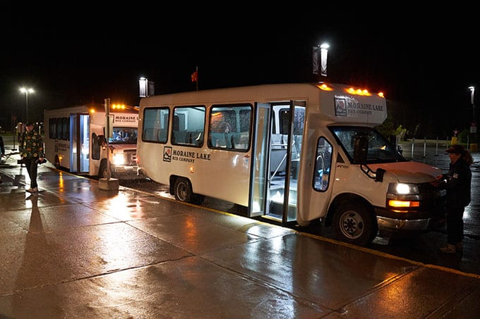 Two white Moraine Lake Bus Company shuttle buses waiting for passengers. It's rainy and dark; they have their lights on.