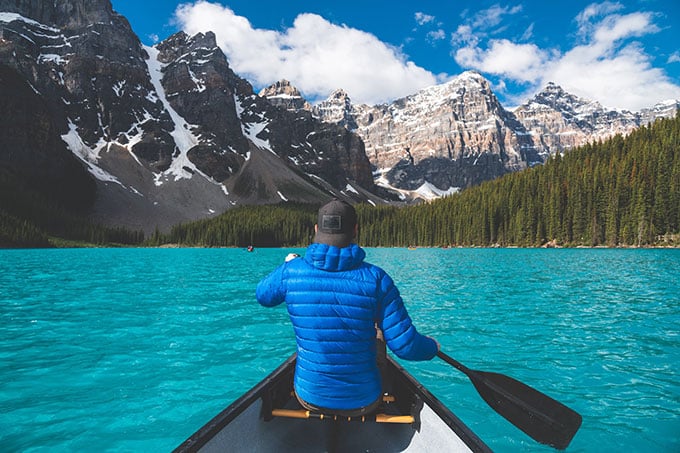 A photo of the back of a person in the front of a canoe paddling across a blue lake. They’re wearing a blue padded jacket.