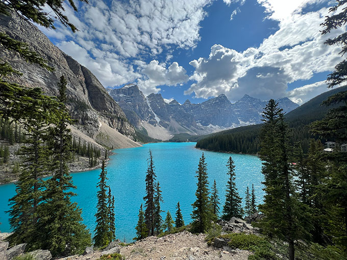 A photo of a vibrant blue lake with trees in the foreground and seven grey peaks of mountains in the background.