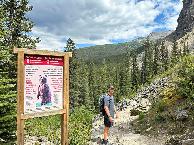 A photo of a white man in a baseball hat and grey t-shirt on a rocky hiking trail. There are trees in the background and a grizzly bear warning sign in the foreground, to the left.